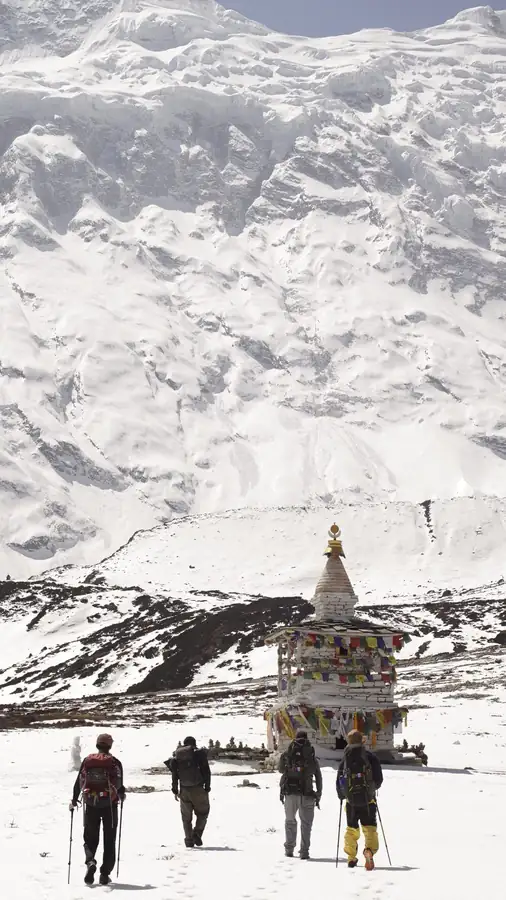 Trekkers walking across snow towards a chorten, with a vast snow wall rising behind