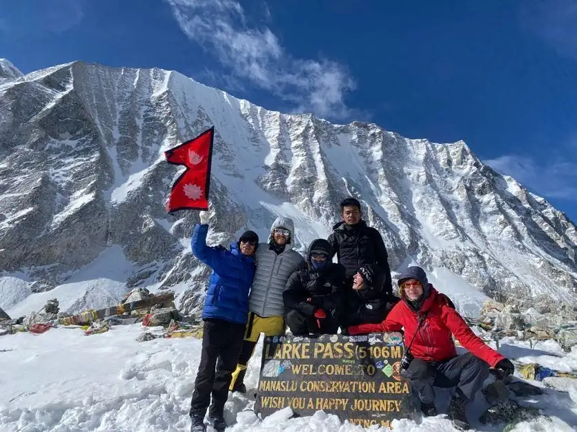 Trekkers holding the Nepal flag at the Larke Pass sign, snow-covered peak behind