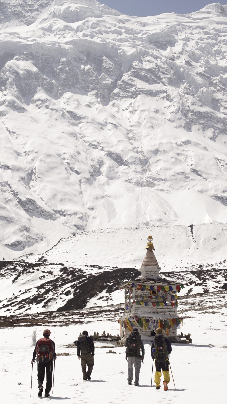 Trekkers walking across snow towards a chorten, with a vast snow wall rising behind