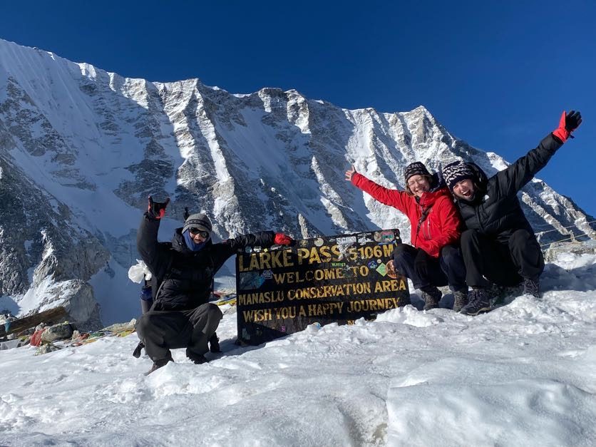 Trekkers with arms raised celebrating at the Larke Pass sign below a wall of snow