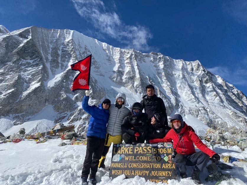 Trekkers holding the Nepal flag at the Larke Pass sign, snow-covered peak behind